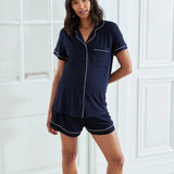 A woman modeling a navy blue short-sleeve pajama set with white piping details, consisting of a button-up shirt and matching shorts, posed in front of white paneled doors.