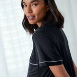 A woman wearing a black short-sleeve top with thin white piping trim on the sleeve and pocket, photographed in a bright indoor setting.