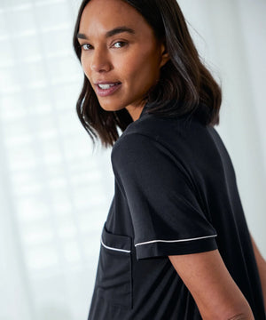 A woman wearing a black short-sleeve top with thin white piping trim on the sleeve and pocket, photographed in a bright indoor setting.