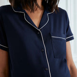 A close-up of a model wearing a navy blue short-sleeve pajama shirt with white piping trim and a button-down front.