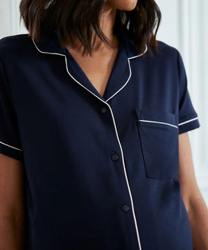 A close-up of a model wearing a navy blue short-sleeve pajama shirt with white piping trim and a button-down front.