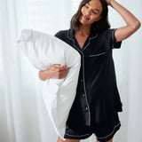 A woman wearing a black short-sleeve pajama set with white piping holds a white pillow in a bright bedroom setting with sheer white curtains.