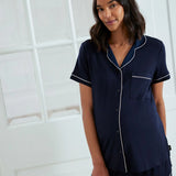 A woman wearing a navy blue short-sleeve pajama set with white piping details, standing in front of a white paneled door.