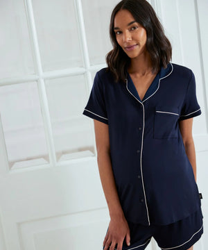 A woman wearing a navy blue short-sleeve pajama set with white piping details, standing in front of a white paneled door.