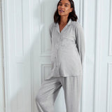 A pregnant woman wearing a light gray heathered two-piece pajama set with a long-sleeve button-front top and matching wide-leg pants, posing against a white paneled wall.