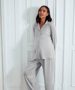 A pregnant woman wearing a light gray heathered two-piece pajama set with a long-sleeve button-front top and matching wide-leg pants, posing against a white paneled wall.