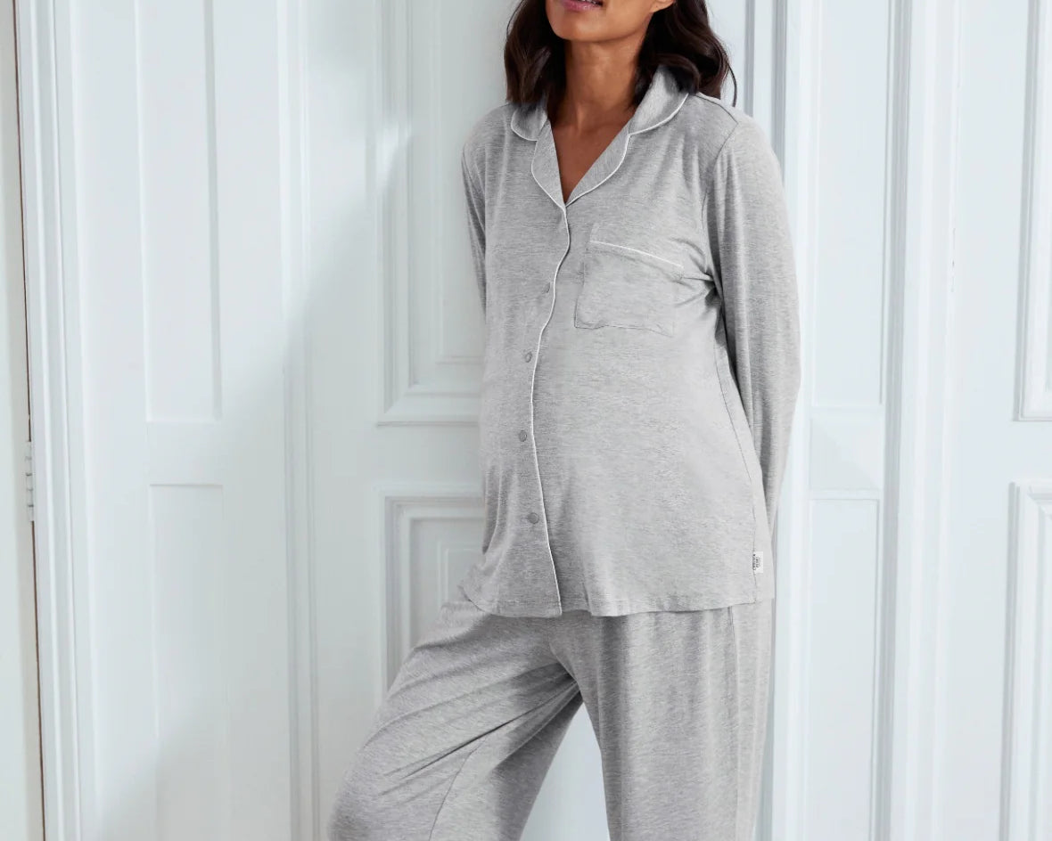 A pregnant woman wearing a light gray heathered two-piece pajama set with a long-sleeve button-front top and matching wide-leg pants, posing against a white paneled wall.