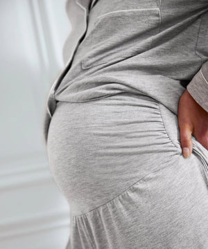 Close-up of a pregnant person wearing a light heather gray maternity dress with a gathered elastic band across the belly and a side pocket, with one hand resting on the hip.