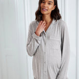 A woman wearing a light gray long-sleeve pajama top with white piping and matching gray pajama pants, standing against a white paneled door.