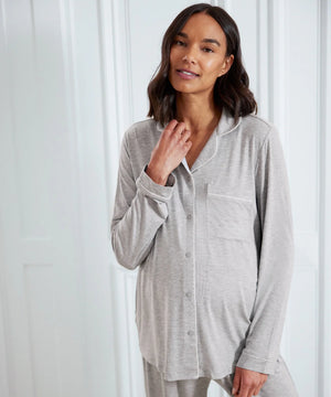 A woman wearing a light gray long-sleeve pajama top with white piping and matching gray pajama pants, standing against a white paneled door.