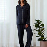 A woman modeling a dark navy blue pajama set with white piping details, standing in a bright room with white curtains and a potted plant.