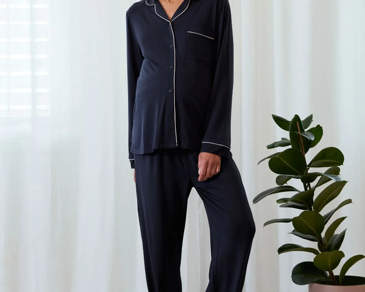 A woman modeling a dark navy blue pajama set with white piping details, standing in a bright room with white curtains and a potted plant.