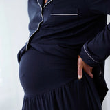 A close-up of a pregnant model wearing a navy blue maternity pajama set with white piping details on a light gray background.
