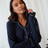A woman wearing a dark navy blue long-sleeve pajama shirt with white piping trim, posed against a soft white curtain background.