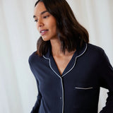 A woman wearing a dark navy blue long-sleeve pajama shirt with white piping trim, set against a soft white curtain background.