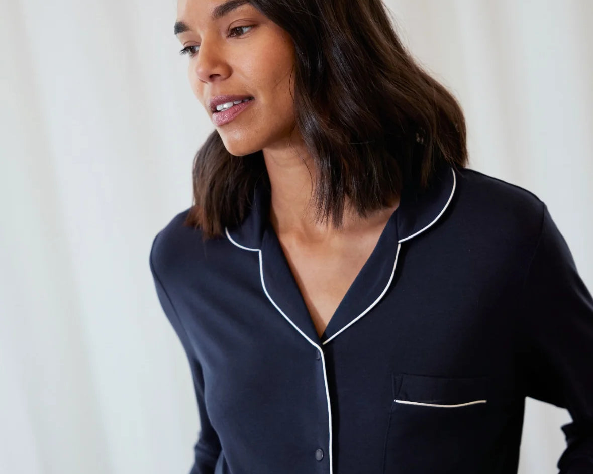 A woman wearing a dark navy blue long-sleeve pajama shirt with white piping trim, set against a soft white curtain background.
