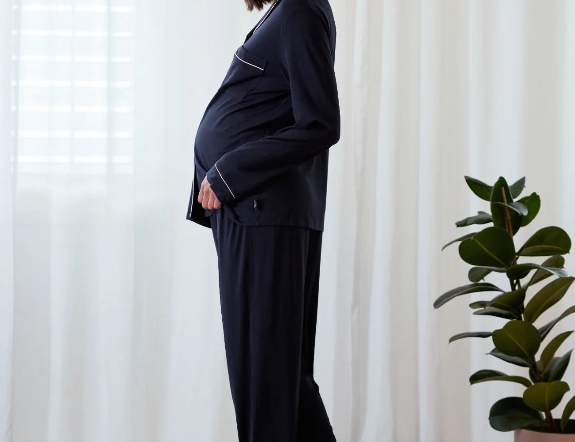 A pregnant woman wearing a dark navy blue pajama set with white piping, standing in a brightly lit room with white curtains and a potted plant.