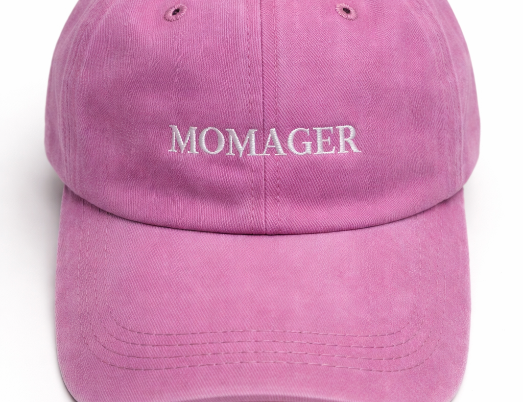 A pink cotton baseball cap with the word 'MOMAGER' embroidered in white across the front, shown against a plain white background.