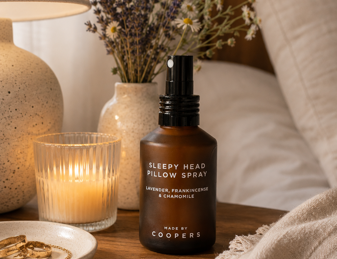 Sleepy Head Pillow Spray on a bedside table with dried lavender and chamomile in a vase, a lit candle and a lamp