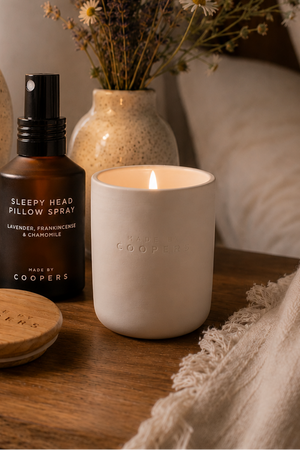 Sleepy Head Pillow Spray beside a lit Made by Coopers candle and its wooden lid on a bedside table