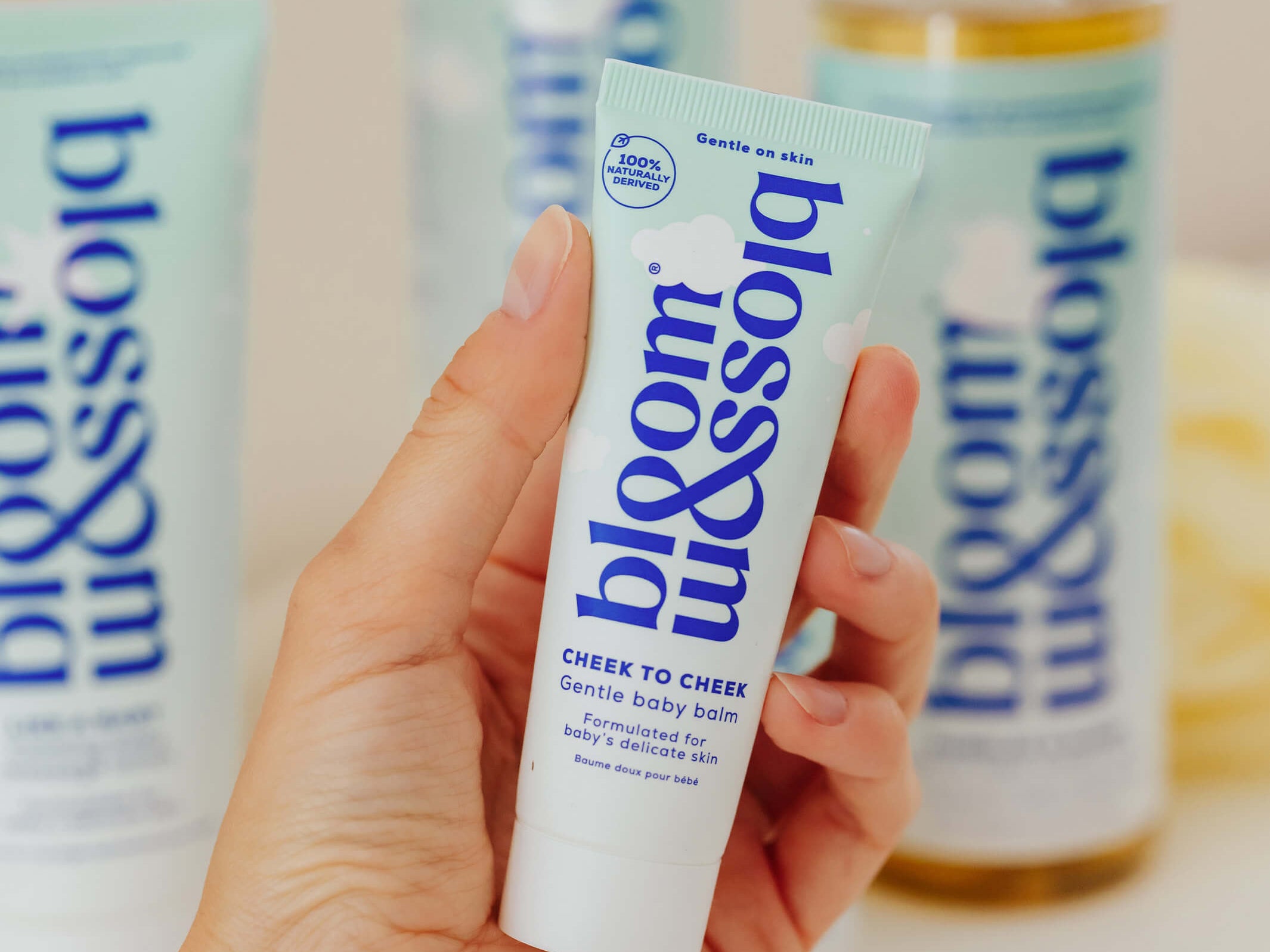 A hand holds a tube of bloom & blossom Cheek to Cheek skin body balm, with other matching body care products blurred in the background.