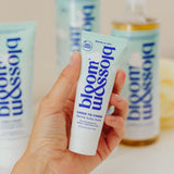 A hand holds a tube of bloom & blossom Cheek to Cheek skin body balm, with other matching body care products blurred in the background.