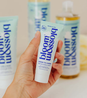 A hand holds a tube of bloom & blossom Cheek to Cheek skin body balm, with other matching body care products blurred in the background.