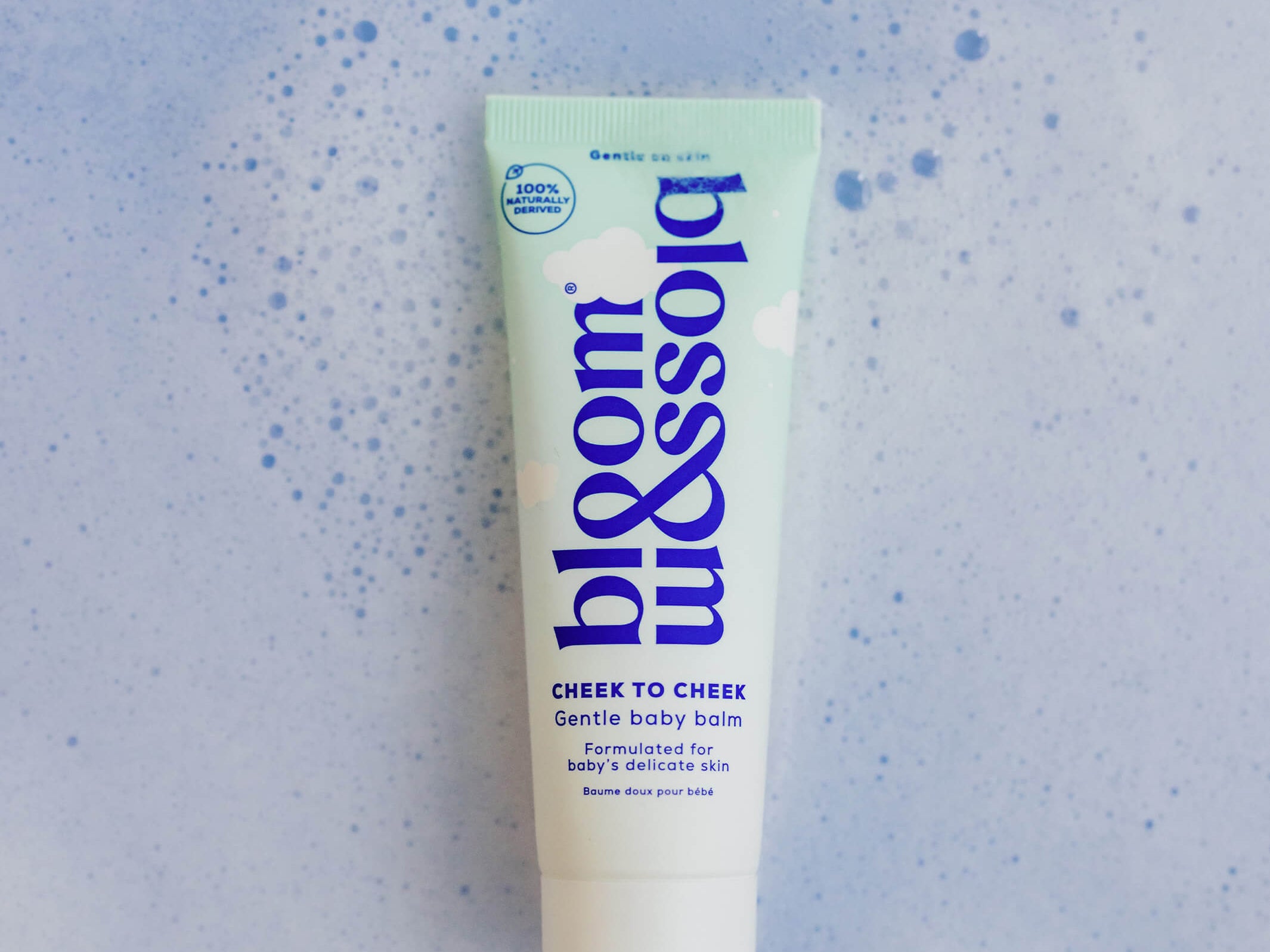 A squeeze tube of bloom & soft Cheek to Cheek Gentle Baby Balm centered on a light blue textured background with subtle speckles.