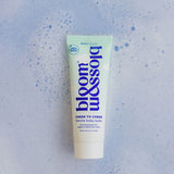 A squeeze tube of bloom & soft Cheek to Cheek Gentle Baby Balm centered on a light blue textured background with subtle speckles.