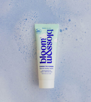 A squeeze tube of bloom & soft Cheek to Cheek Gentle Baby Balm centered on a light blue textured background with subtle speckles.