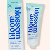 Blossom Cheek to Cheek gentle baby balm shown as a light blue squeeze tube next to its matching retail box on a pale peach background.