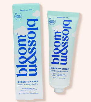 Blossom Cheek to Cheek gentle baby balm shown as a light blue squeeze tube next to its matching retail box on a pale peach background.