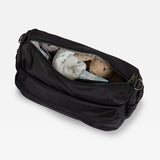 A black nylon diaper bag shown open, revealing a white diaper and a plush teddy bear inside, set against a plain light gray background.