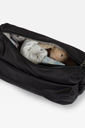 A black nylon diaper bag shown open, revealing a white diaper and a plush teddy bear inside, set against a plain light gray background.