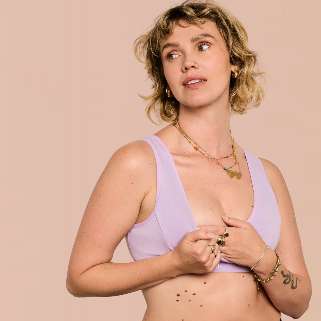 A woman with short wavy blonde hair wearing a light purple/lavender bra, posing against a solid pale peach background. She is wearing layered gold necklaces, rings, and bracelets.