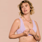 A woman with short wavy blonde hair wearing a light purple/lavender bra, posing against a solid pale peach background. She is wearing layered gold necklaces, rings, and bracelets.