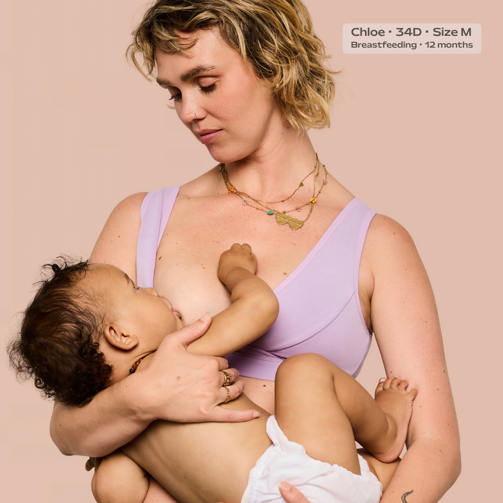 A woman in a light purple nursing bra holds and breastfeeds a baby against a solid pale pink background. A text overlay in the top right reads 'Chloe • 34D • Size M • Breastfeeding • 12 months'.