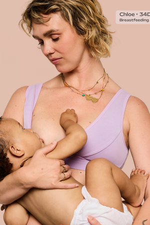 A woman in a light purple nursing bra holds and breastfeeds a baby against a solid pale pink background. A text overlay in the top right reads 'Chloe • 34D • Size M • Breastfeeding • 12 months'.