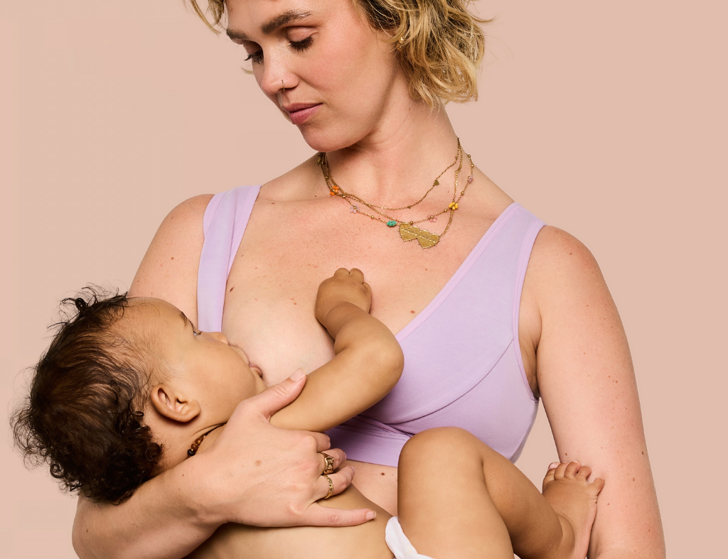 A woman in a light purple nursing bra holds and breastfeeds a baby against a solid pale pink background. A text overlay in the top right reads 'Chloe • 34D • Size M • Breastfeeding • 12 months'.