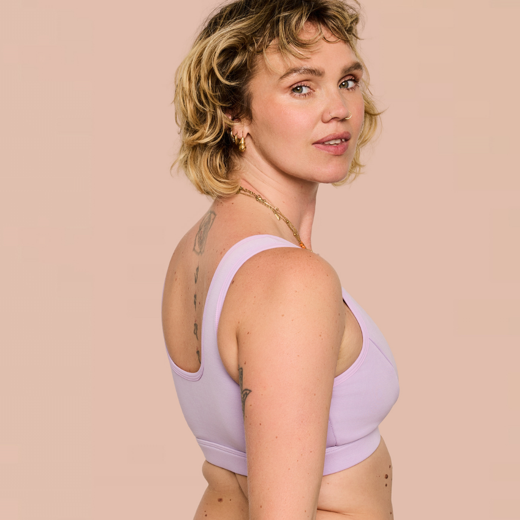A woman with short blonde hair wearing a light purple/lavender sports bra, shown from a side-back angle against a solid pale pink background.