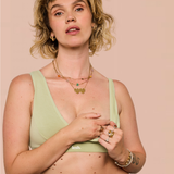 A woman wearing a light sage green bralette with a small white 'kesh.' logo on the band, styled with layered gold necklaces and rings against a solid pale pink background.