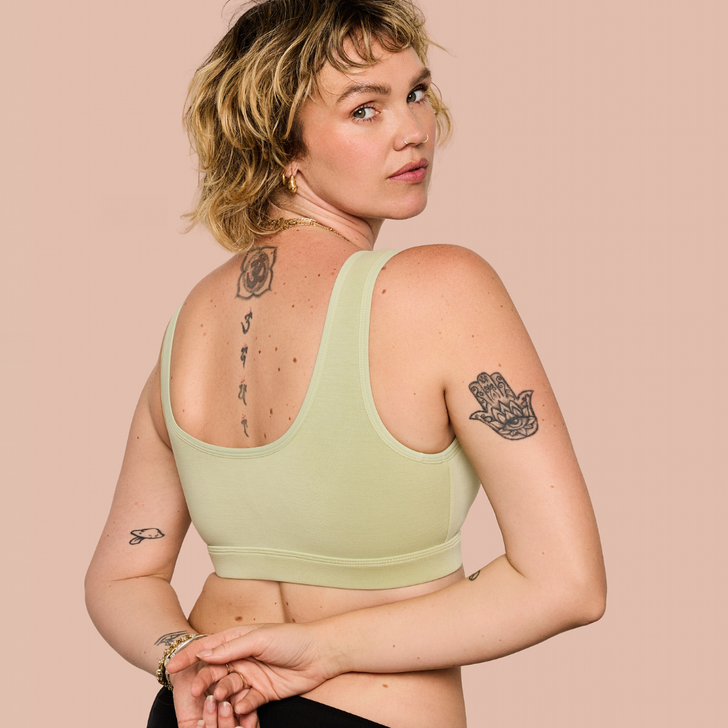 A woman with short blonde hair is shown from the back wearing a light sage green sports bra against a solid pale pink background.