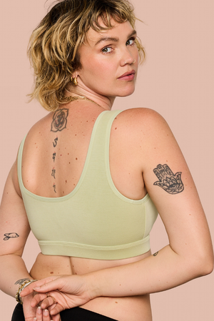 A woman with short blonde hair is shown from the back wearing a light sage green sports bra against a solid pale pink background.