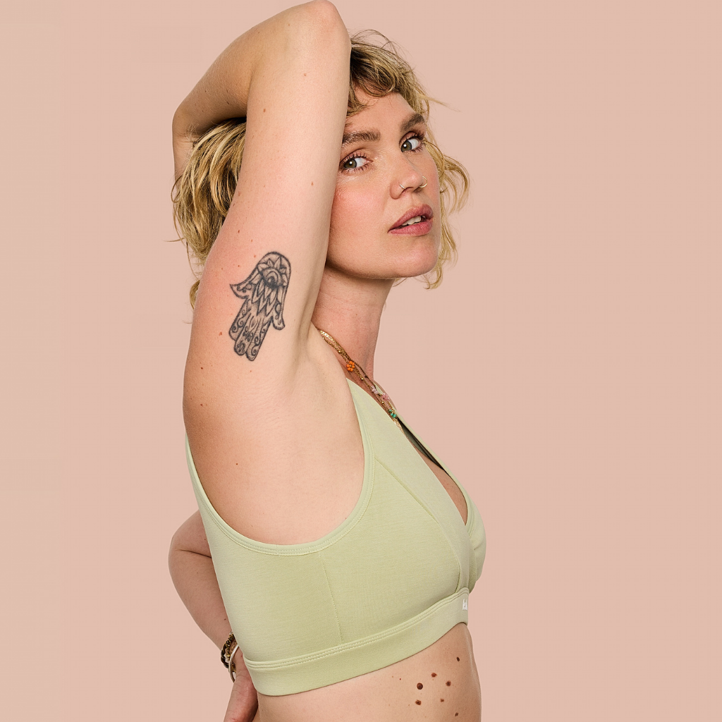 A woman wearing a light sage green sports bra, posing with one arm raised against a solid pale pink background.