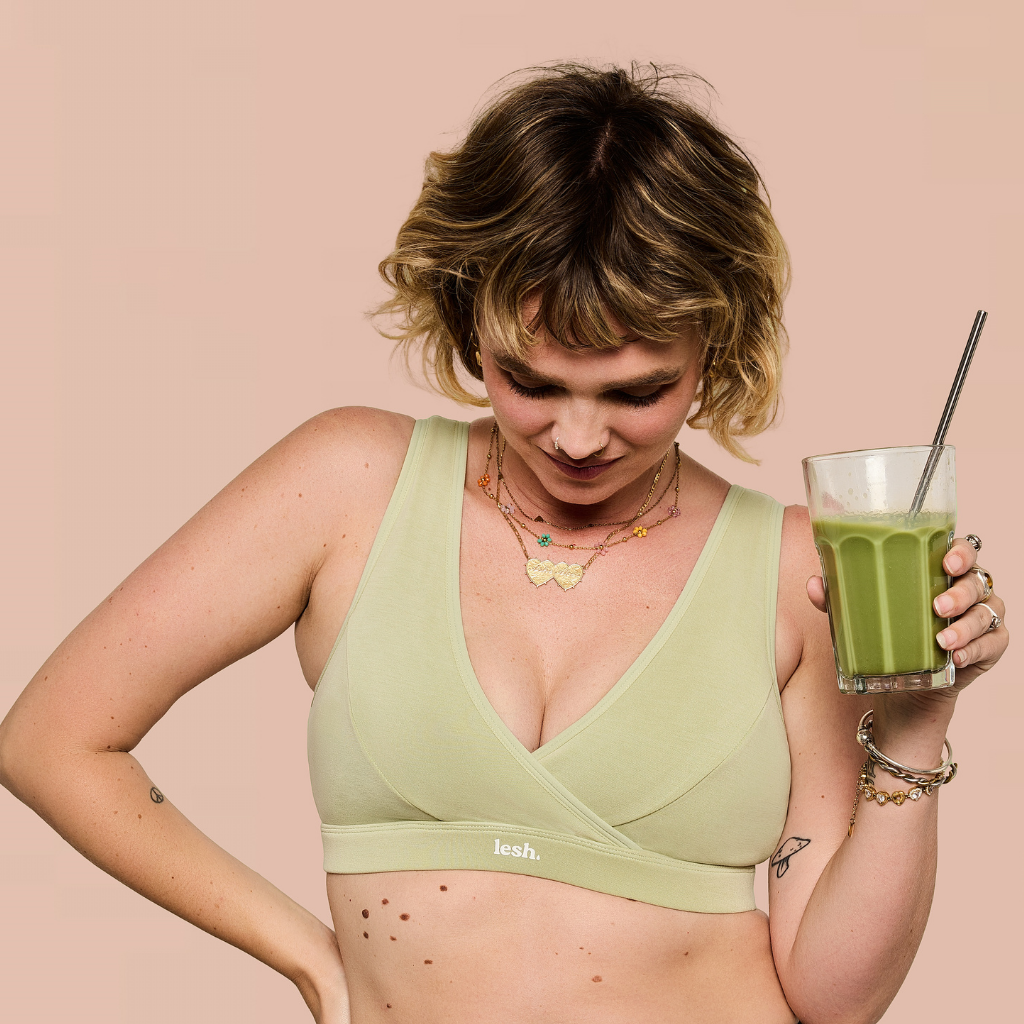 A woman wearing a light sage green bra from the brand leash., holding a glass of green juice against a solid pale pink background.