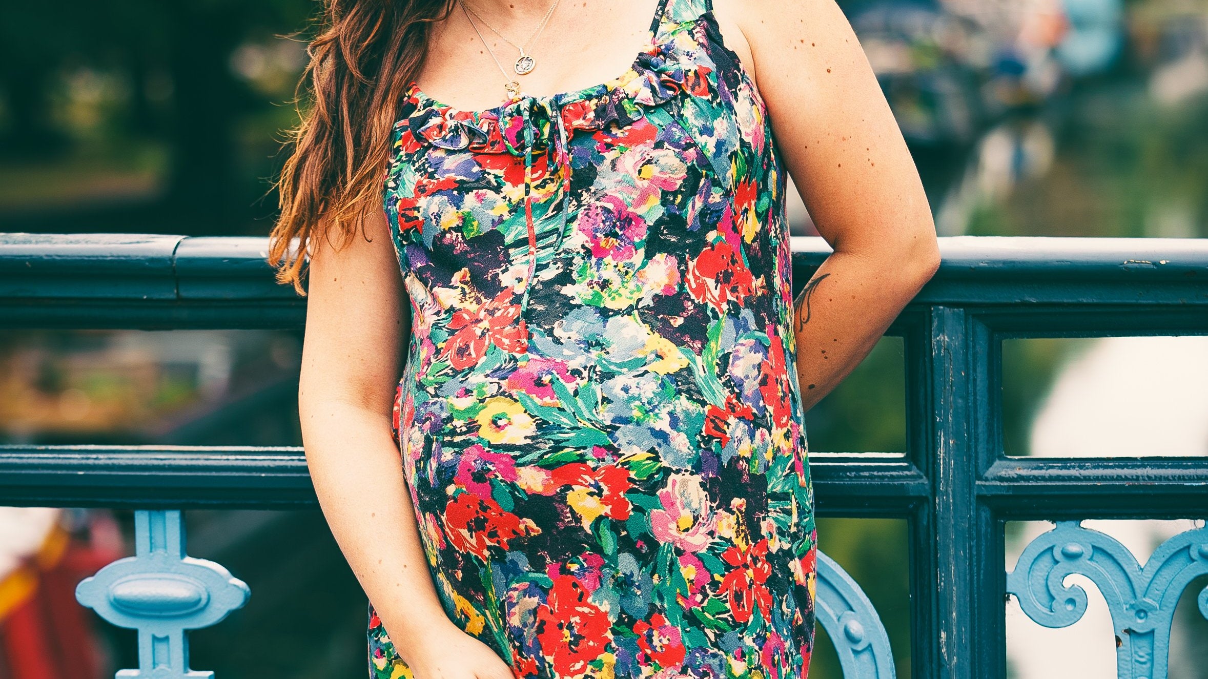 A pregnant woman with long brown hair stands on a blue metal bridge outdoors, wearing a sleeveless floral maternity dress with a black background and red, yellow, and white flowers.