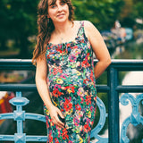 A pregnant woman with long brown hair stands on a blue metal bridge outdoors, wearing a sleeveless floral maternity dress with a black background and red, yellow, and white flowers.