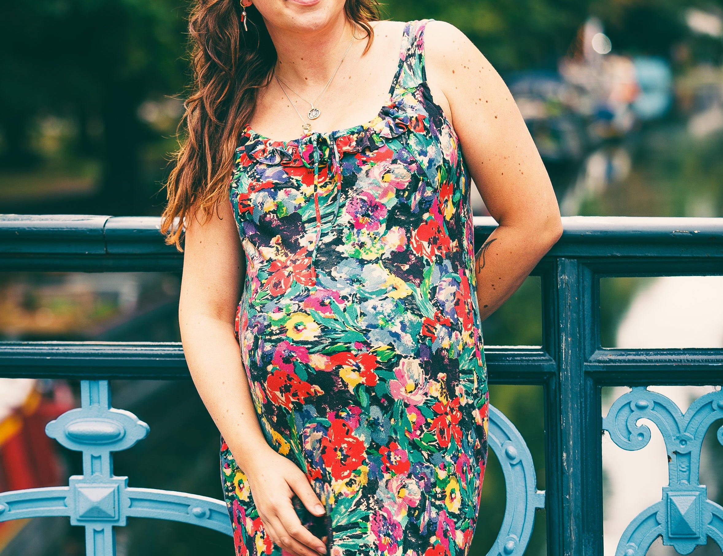A pregnant woman with long brown hair stands on a blue metal bridge outdoors, wearing a sleeveless floral maternity dress with a black background and red, yellow, and white flowers.