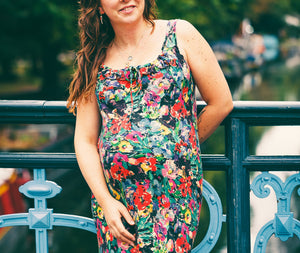 A pregnant woman with long brown hair stands on a blue metal bridge outdoors, wearing a sleeveless floral maternity dress with a black background and red, yellow, and white flowers.