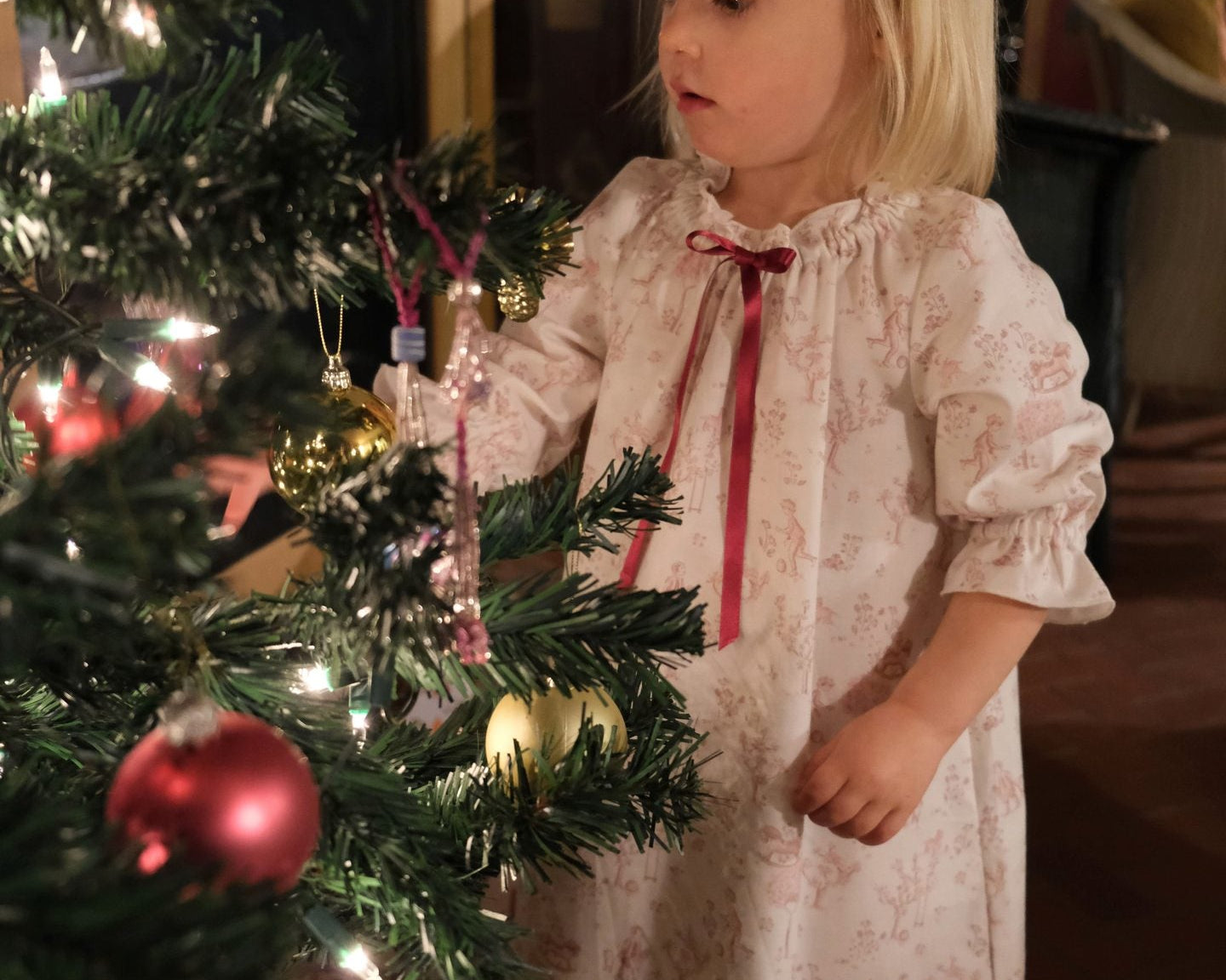 Girls' Nightie in Cordelia Toile, child in the pink toile print with red ribbon tie looking at baubles on a Christmas tree
